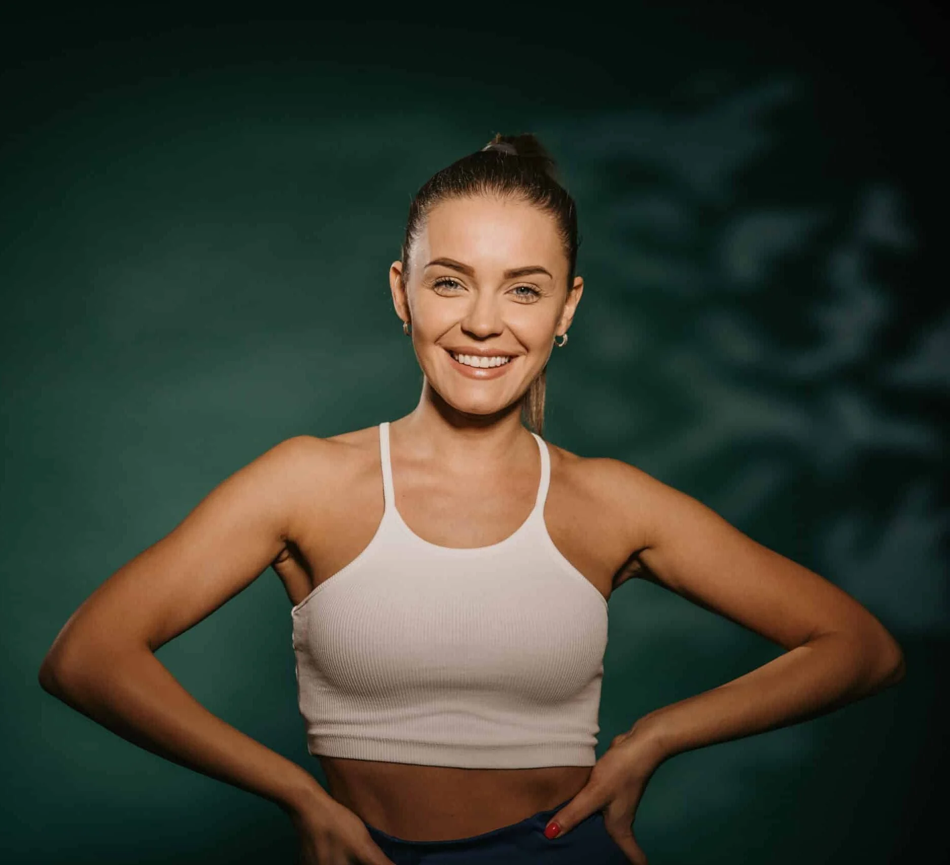 Smiling young woman standing in studio while looking at camera with arms on hips